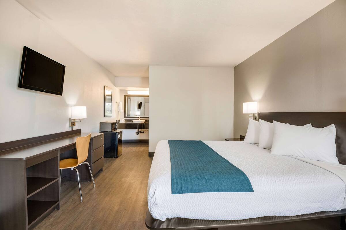 A hotel room with a bed, a desk, a chair, a TV, and a bathroom in the background.