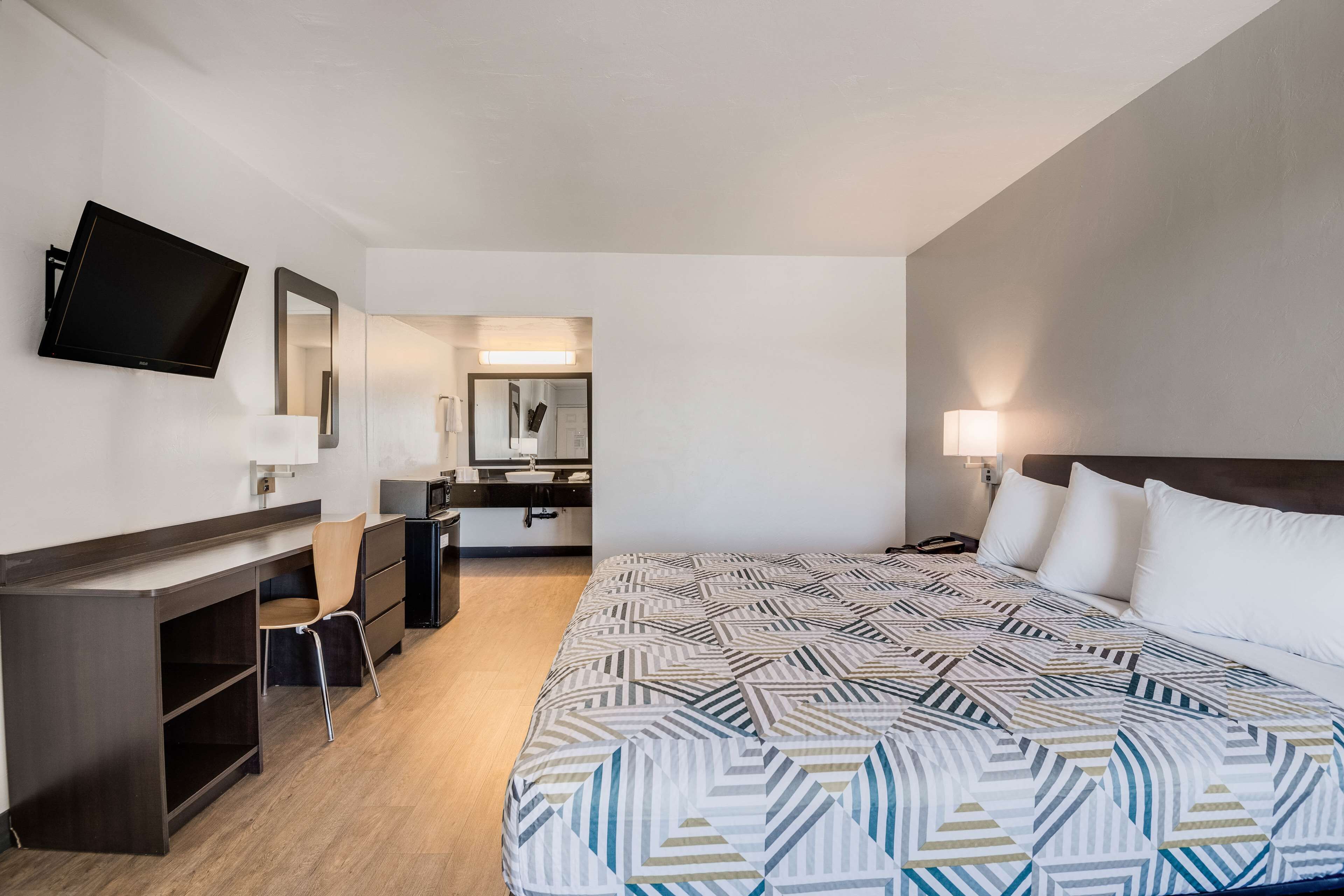 A hotel room with a bed, a desk, a chair, a TV, a mirror, and a bathroom visible in the background.