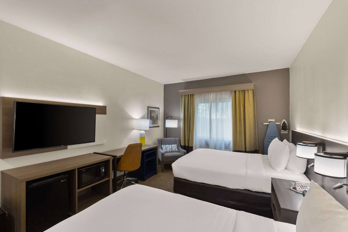 A hotel room with two beds, a TV, a desk with a microwave, a chair, and a window with curtains.