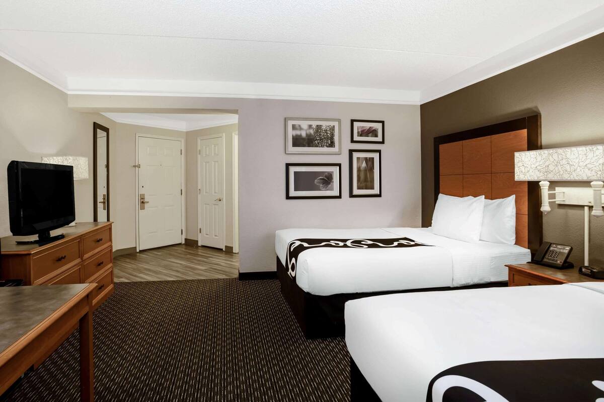 A hotel room with two beds, a desk, a television, and framed pictures on the wall.
