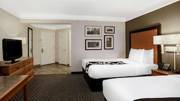 A hotel room with two beds, a desk, a television, and framed pictures on the wall.