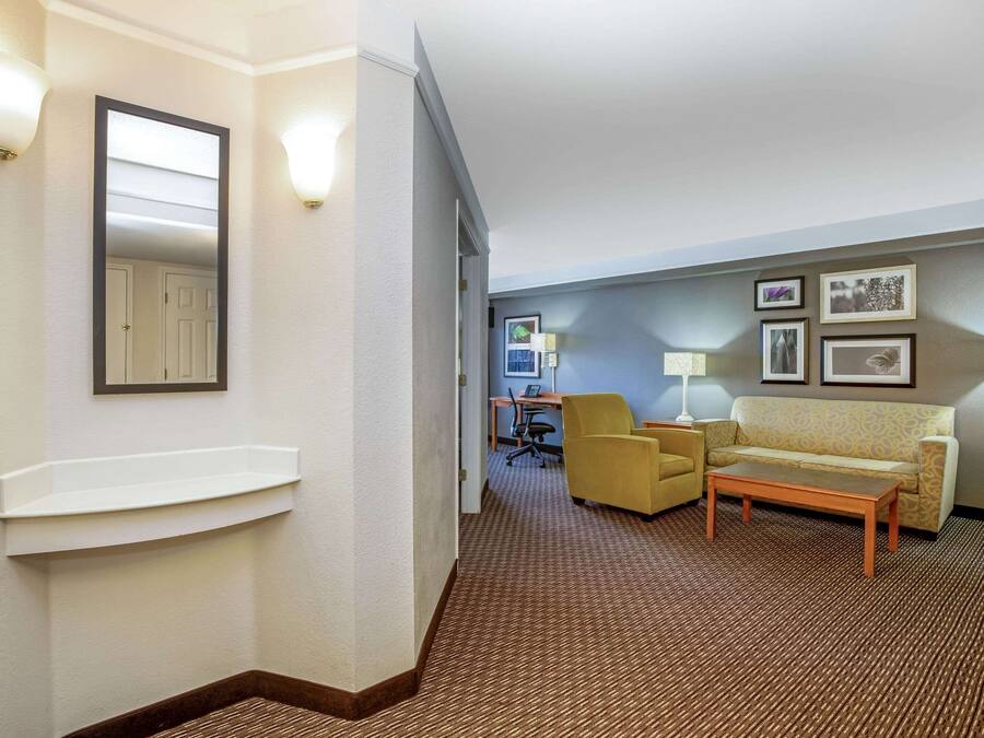 A hotel room with a brown carpet, a sofa, a coffee table, a desk with a chair, and wall-mounted lights.