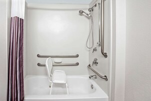 Baño con bañera blanca, ducha con cabezal de ducha manual, taburete blanco y barras de apoyo.
