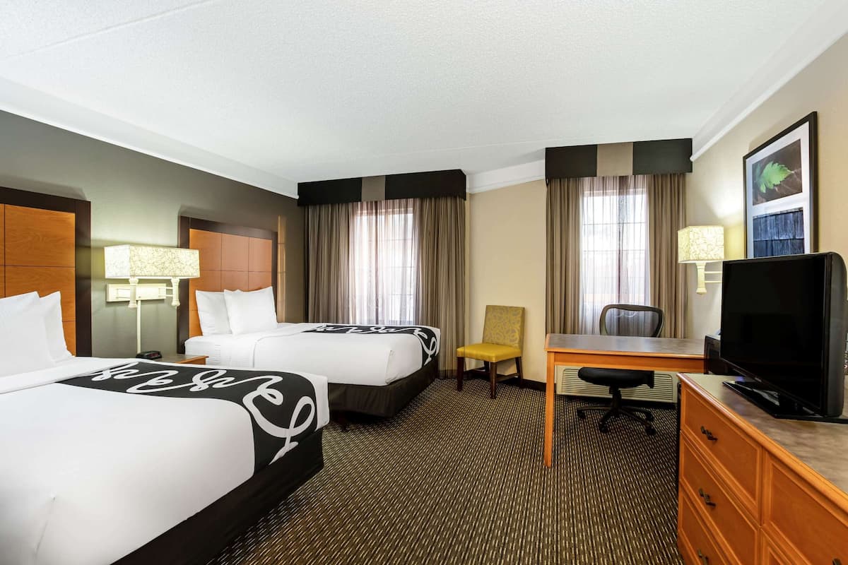 A hotel room with two beds, a desk, a chair, a TV, and a lamp.
