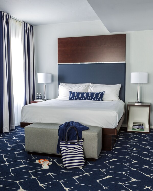 A hotel room with a large bed, a bedside table with a lamp, a nightstand with a lamp, a window with curtains, and a patterned carpet.