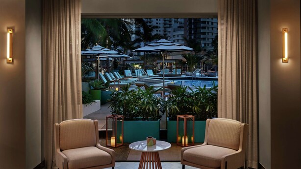 A hotel room with a view of a pool area, two armchairs, a round table, and a potted plant. - The Gates Hotel South Beach (Miami Beach)