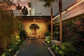 La entrada del The Gates Hotel, con un camino bordeado de plantas y un gran árbol en el centro.