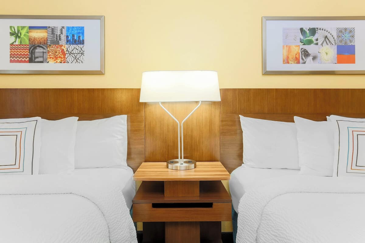 Habitación de hotel con dos camas, un cabecero de madera, una mesita de noche con lámpara y dos cuadros enmarcados en la pared.
