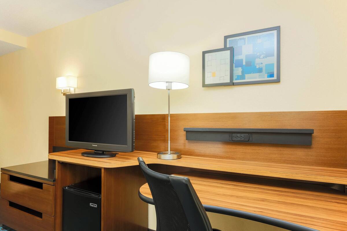 Habitación de hotel con escritorio de madera, televisor de pantalla plana, lámpara, silla y refrigerador pequeño.