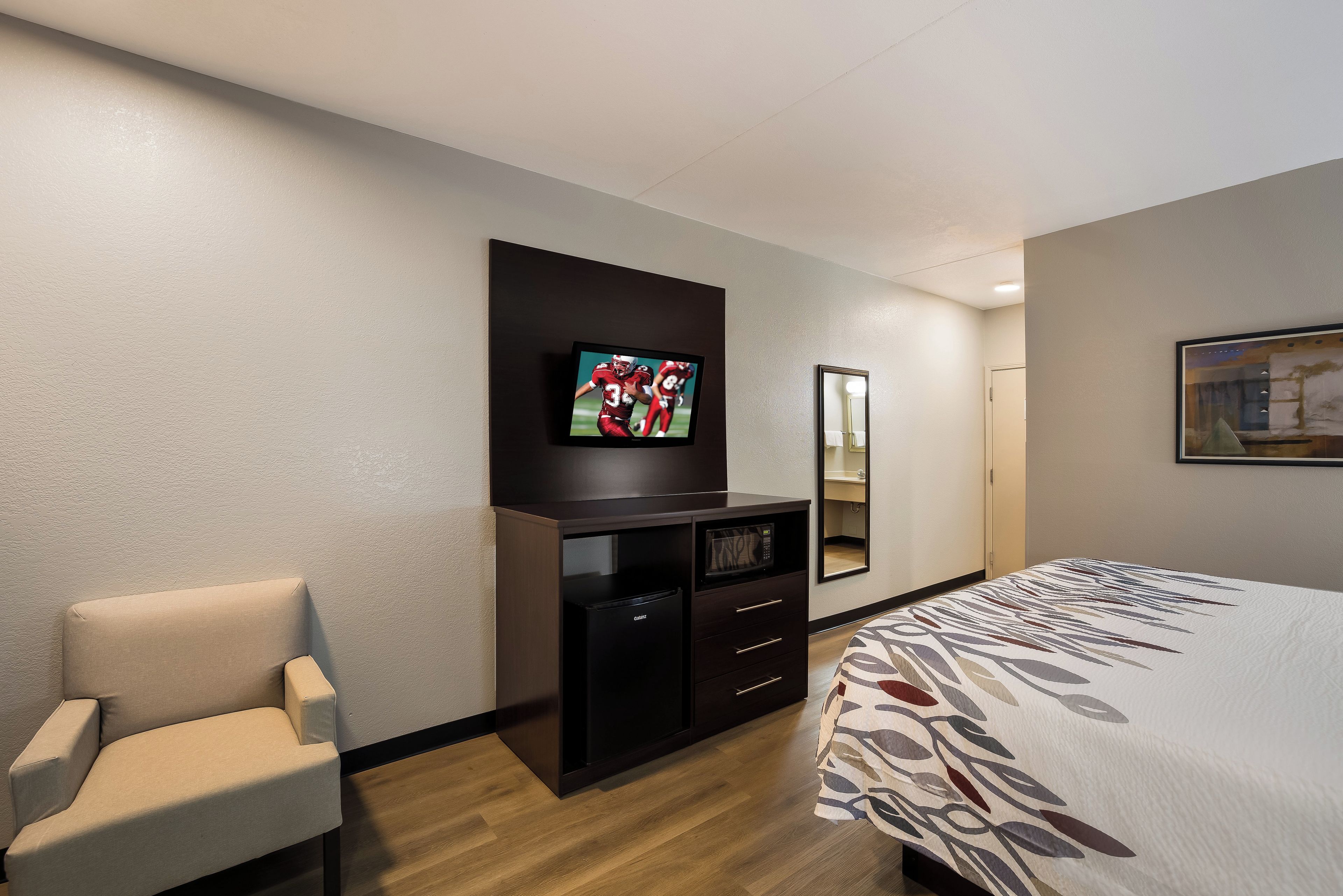A hotel room with a bed, a TV mounted on the wall, a desk with a microwave, a chair, and a mirror.