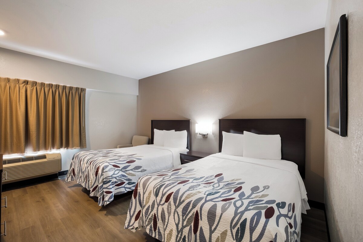 A hotel room with two beds, a heater, a window with curtains, and a wall-mounted light.