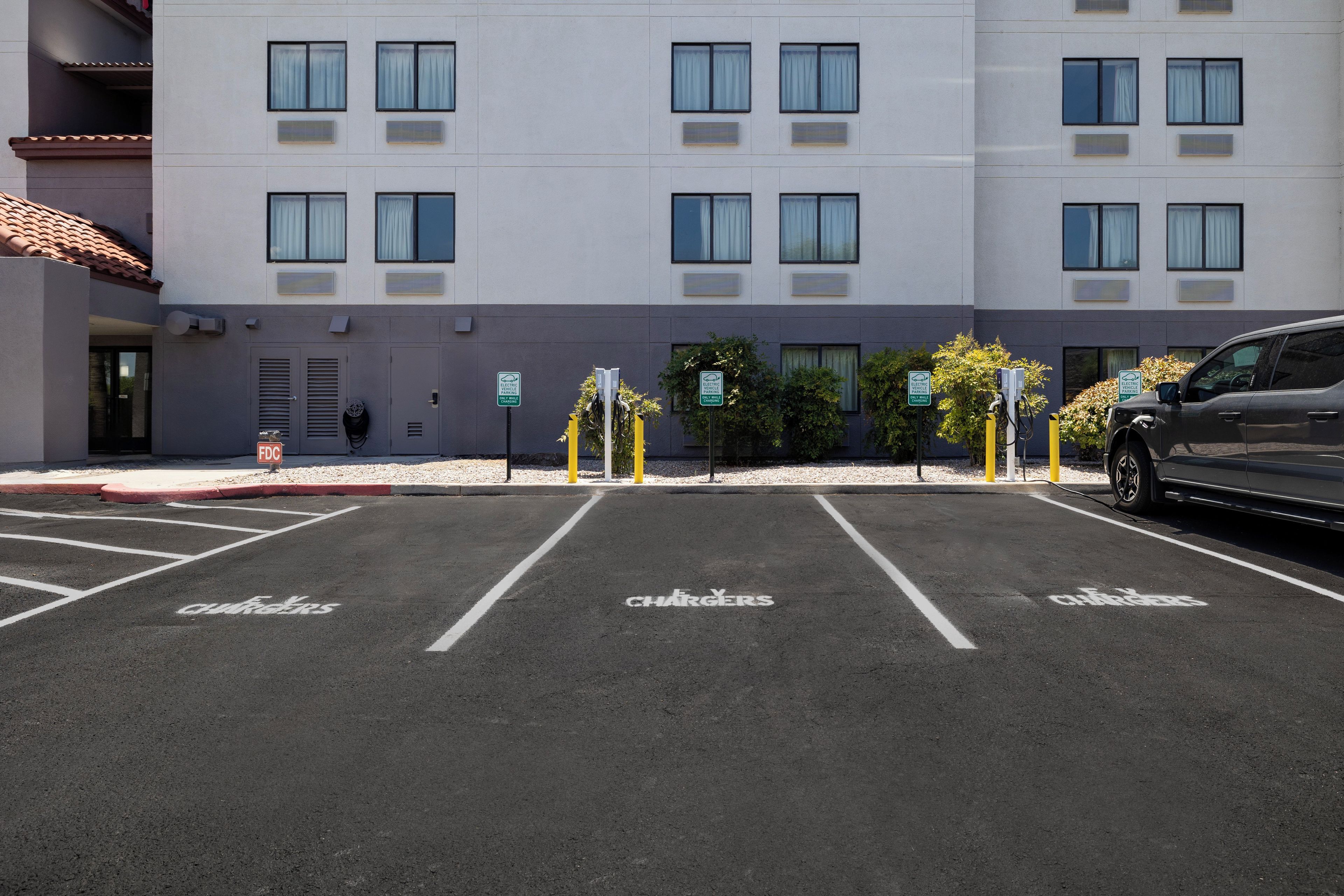 Un parking équipé de bornes de recharge pour véhicules électriques, avec des emplacements de stationnement clairement délimités, et un bâtiment doté de nombreuses fenêtres et de climatiseurs.