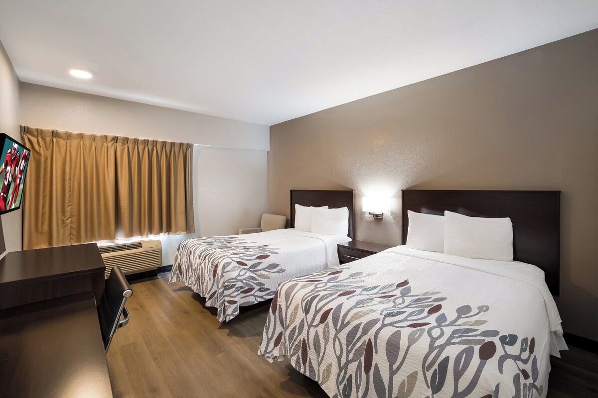A hotel room with two beds, a desk, a chair, a TV, and a window with curtains.