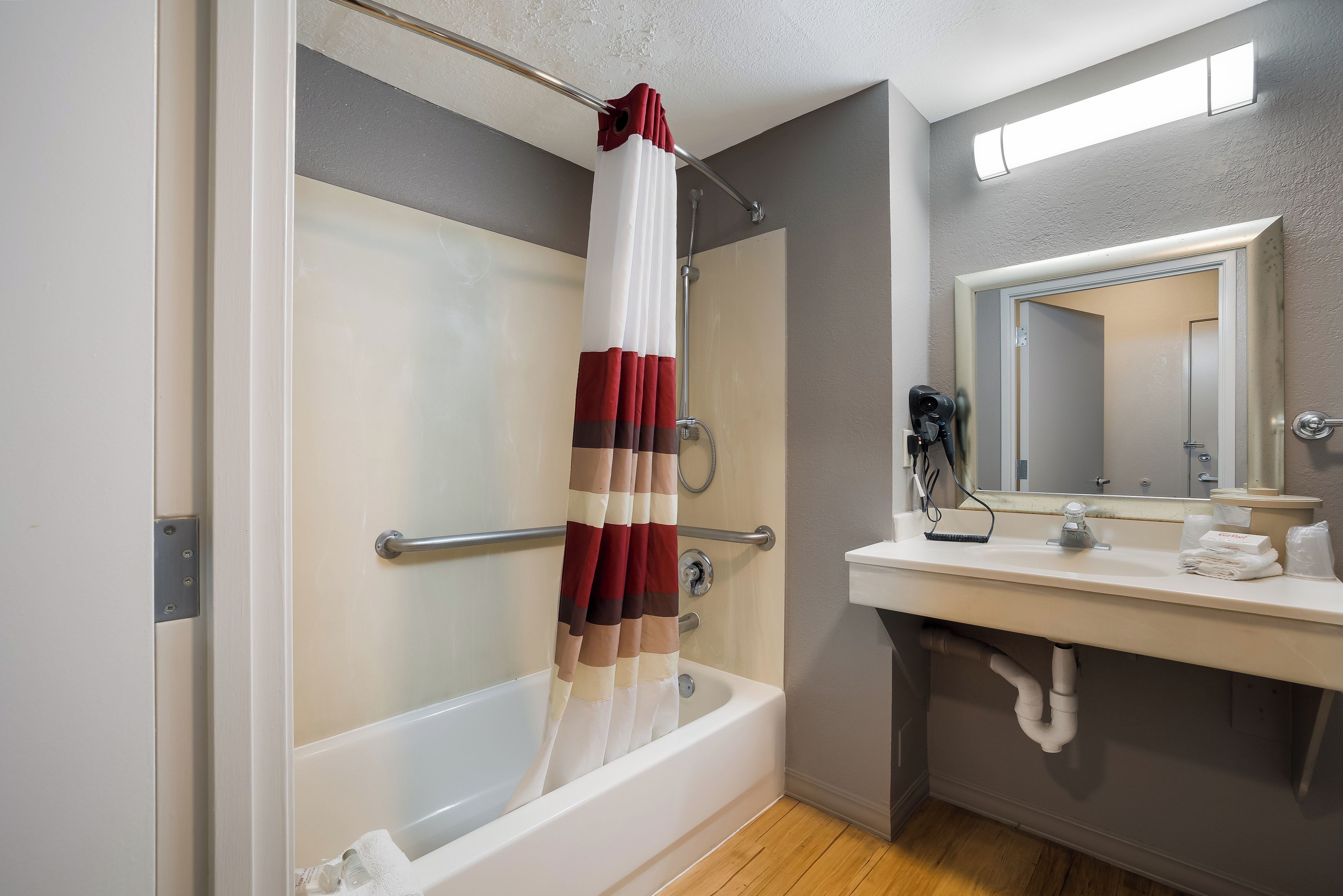 A bathroom with a bathtub, a shower curtain, a handrail, a towel rack, a sink with a mirror, and a towel dispenser.