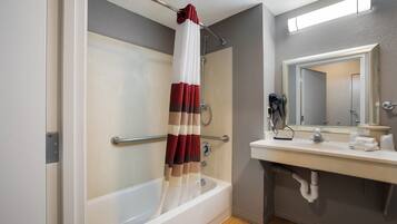 A bathroom with a bathtub, a shower curtain, a handrail, a towel rack, a sink with a mirror, and a towel dispenser.