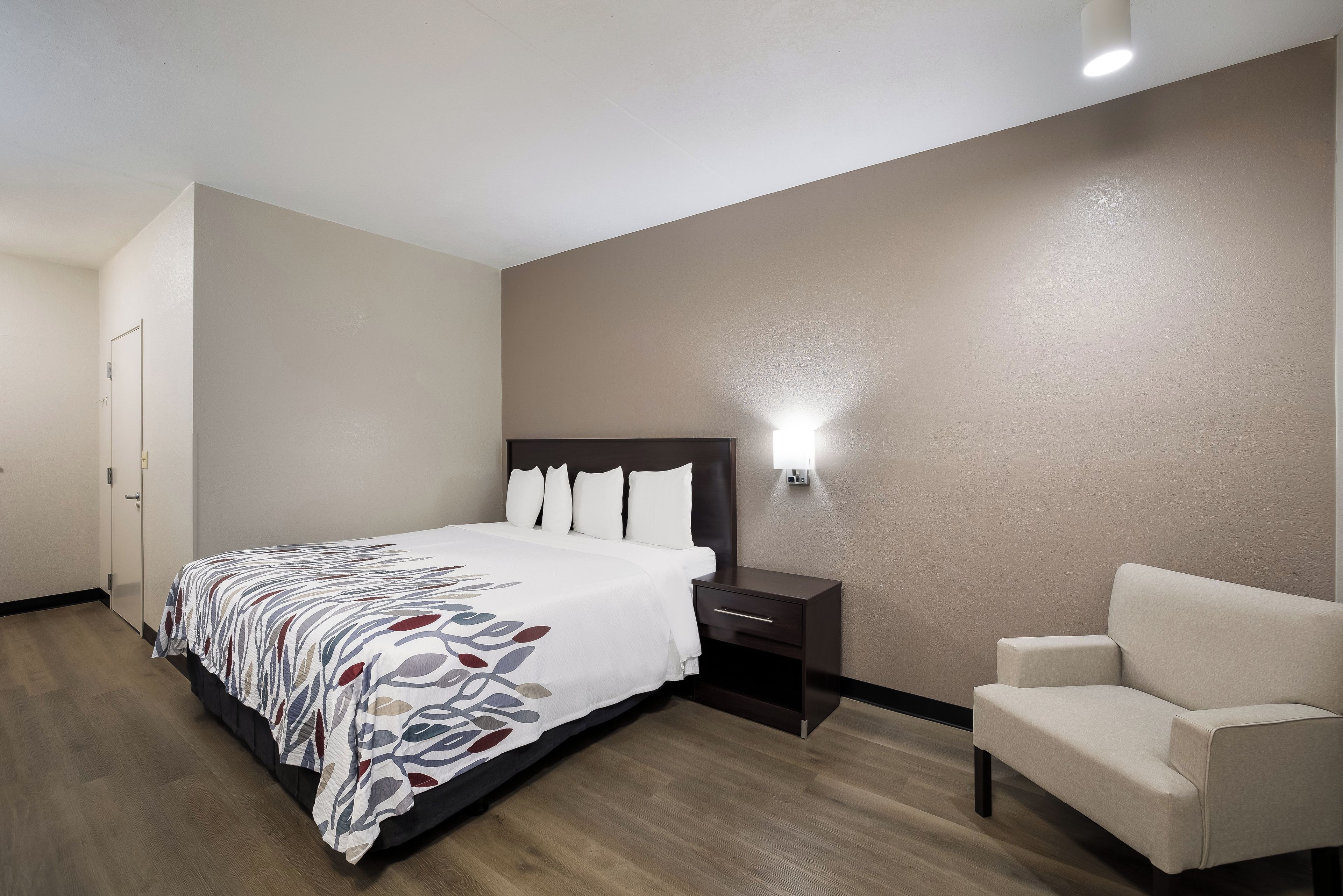 A hotel room with a bed, a nightstand, a chair, and a wall-mounted light.