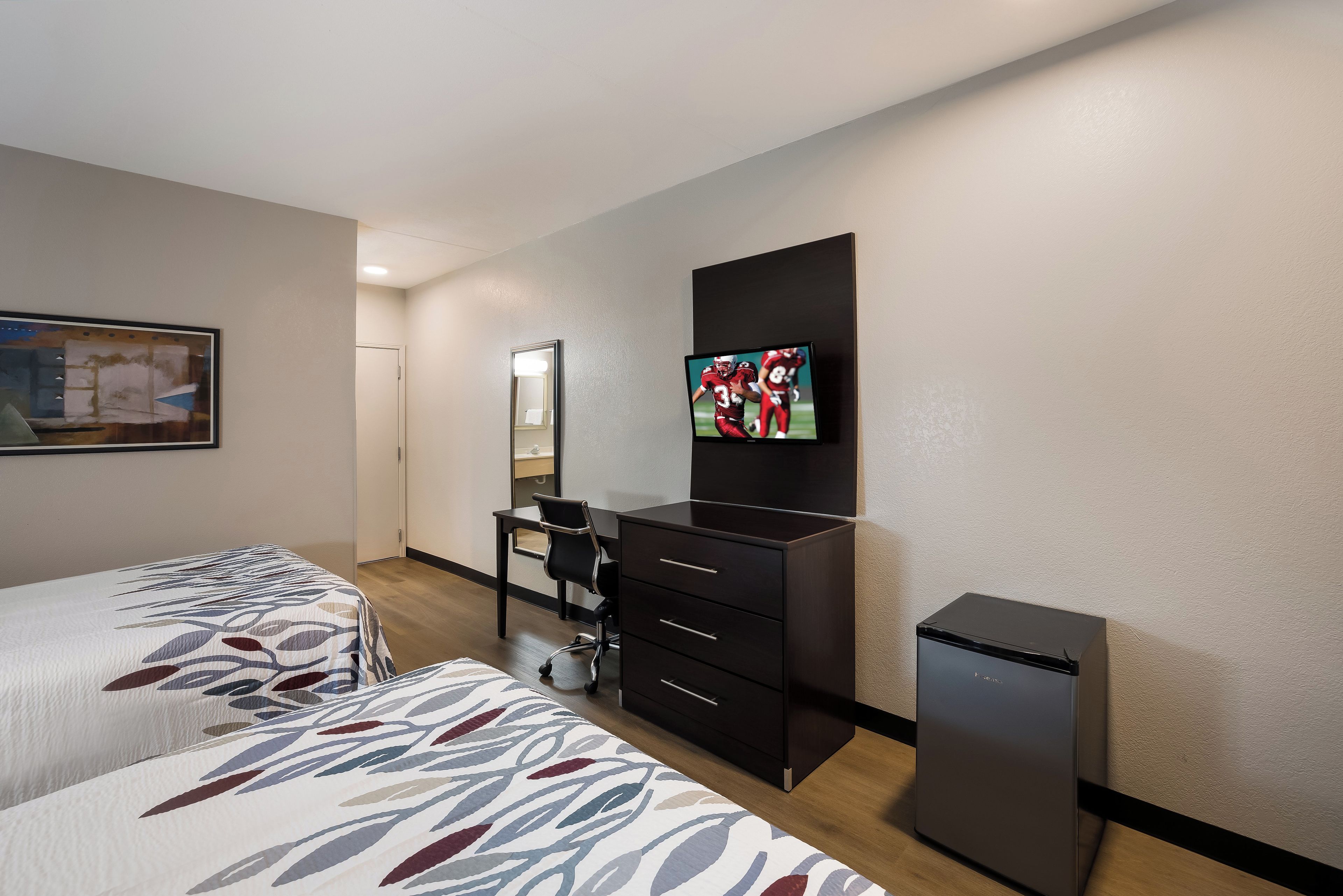 Quarto de hotel com duas camas, uma escrivaninha, uma cadeira, televisão e um pequeno frigorífico.
