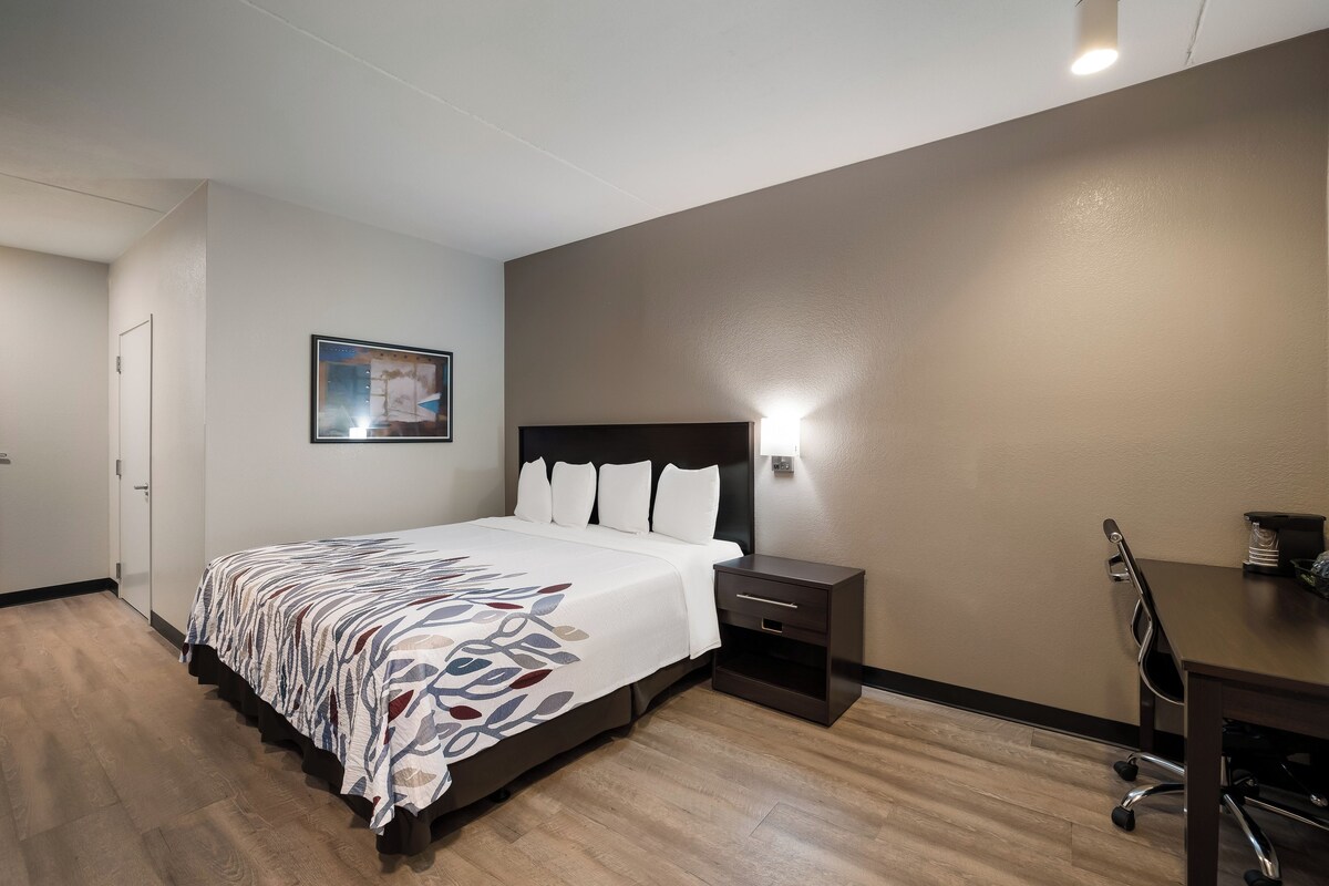 A hotel room with a bed, bedside table, desk, chair, and a painting on the wall.