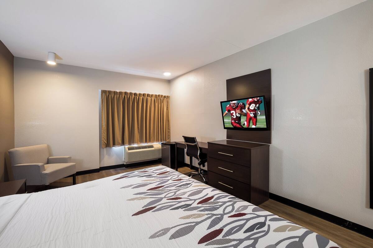 Una habitación de hotel con cama, escritorio, silla, televisor y ventana con cortinas.