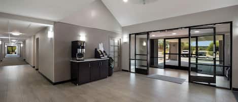 A modern hotel lobby with a reception desk, a coffee machine, and a large sliding glass door leading to an outdoor area.