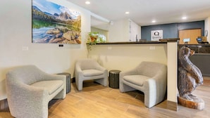 Un área de recepción con tres sillones grises, un mostrador de madera, un letrero que dice 'Quality' y un cuadro de paisaje grande.