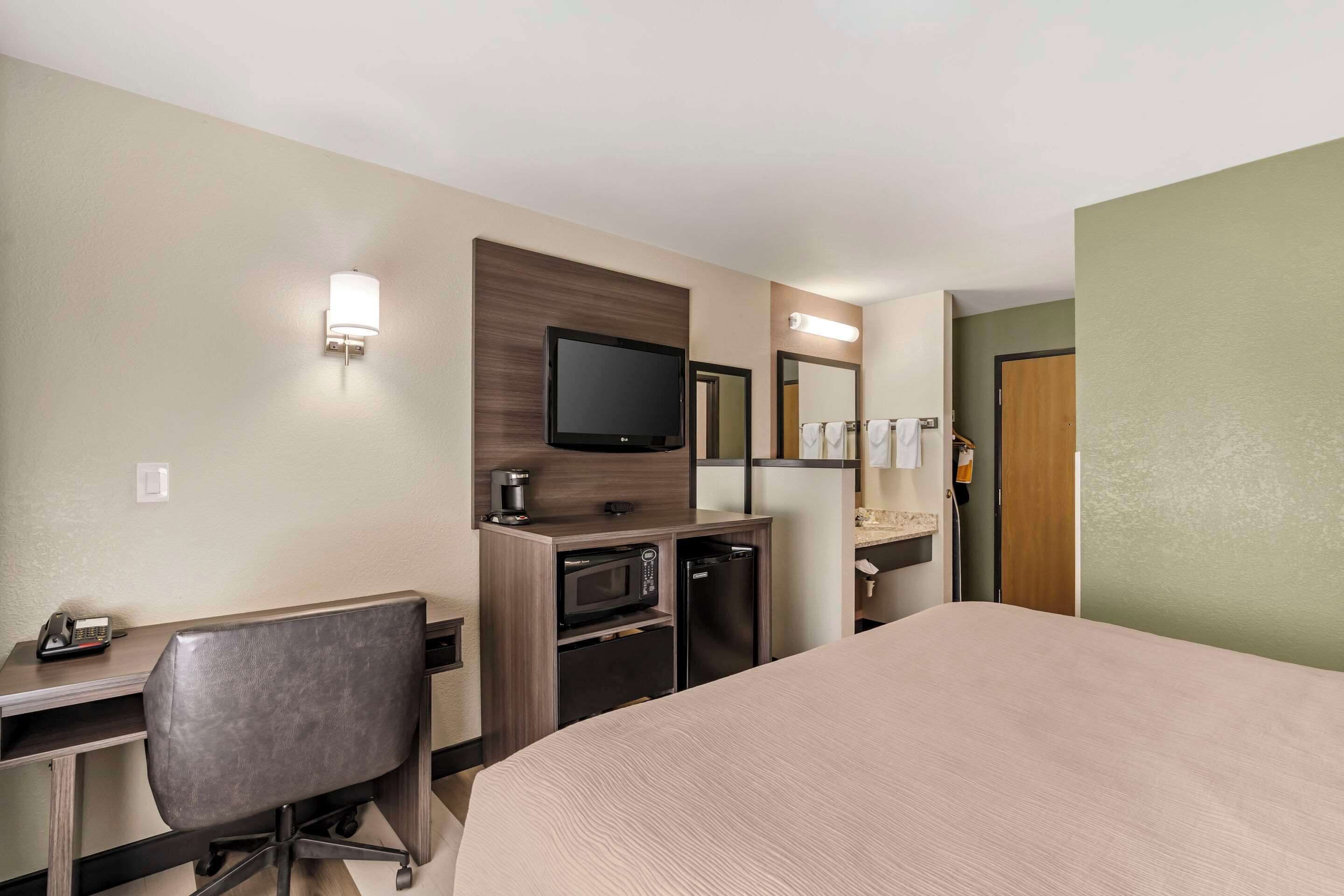 A hotel room with a bed, a desk with a chair, a microwave, a coffee maker, a television, and a bathroom with a mirror and towel rack.
