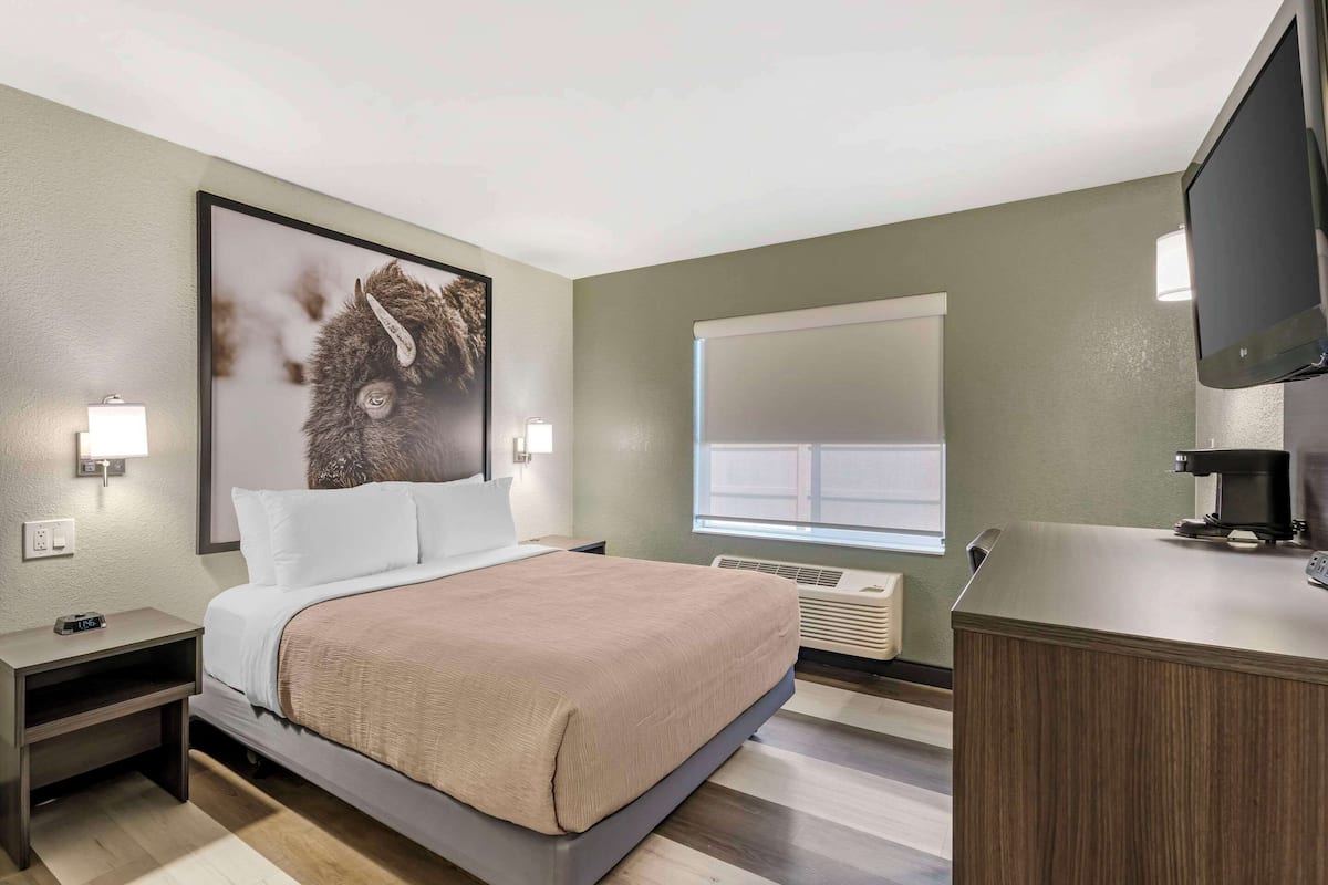 Ein Hotelzimmer mit Bett, Nachttischen, einem Flachbildfernseher, einer Kaffeemaschine und einem großen Fenster mit Jalousien.