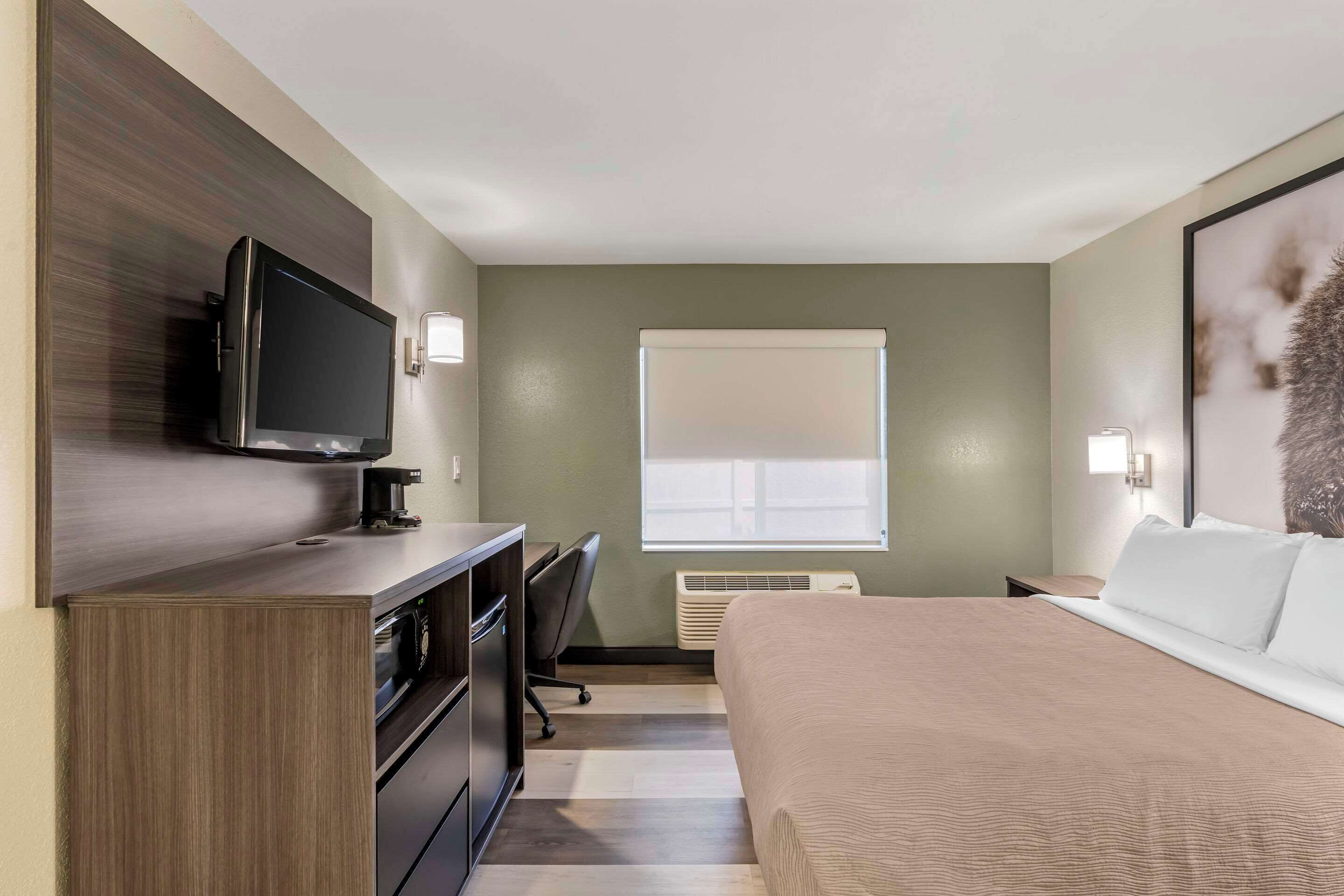 A hotel room with a bed, a desk with a chair, a television, and a window with blinds.