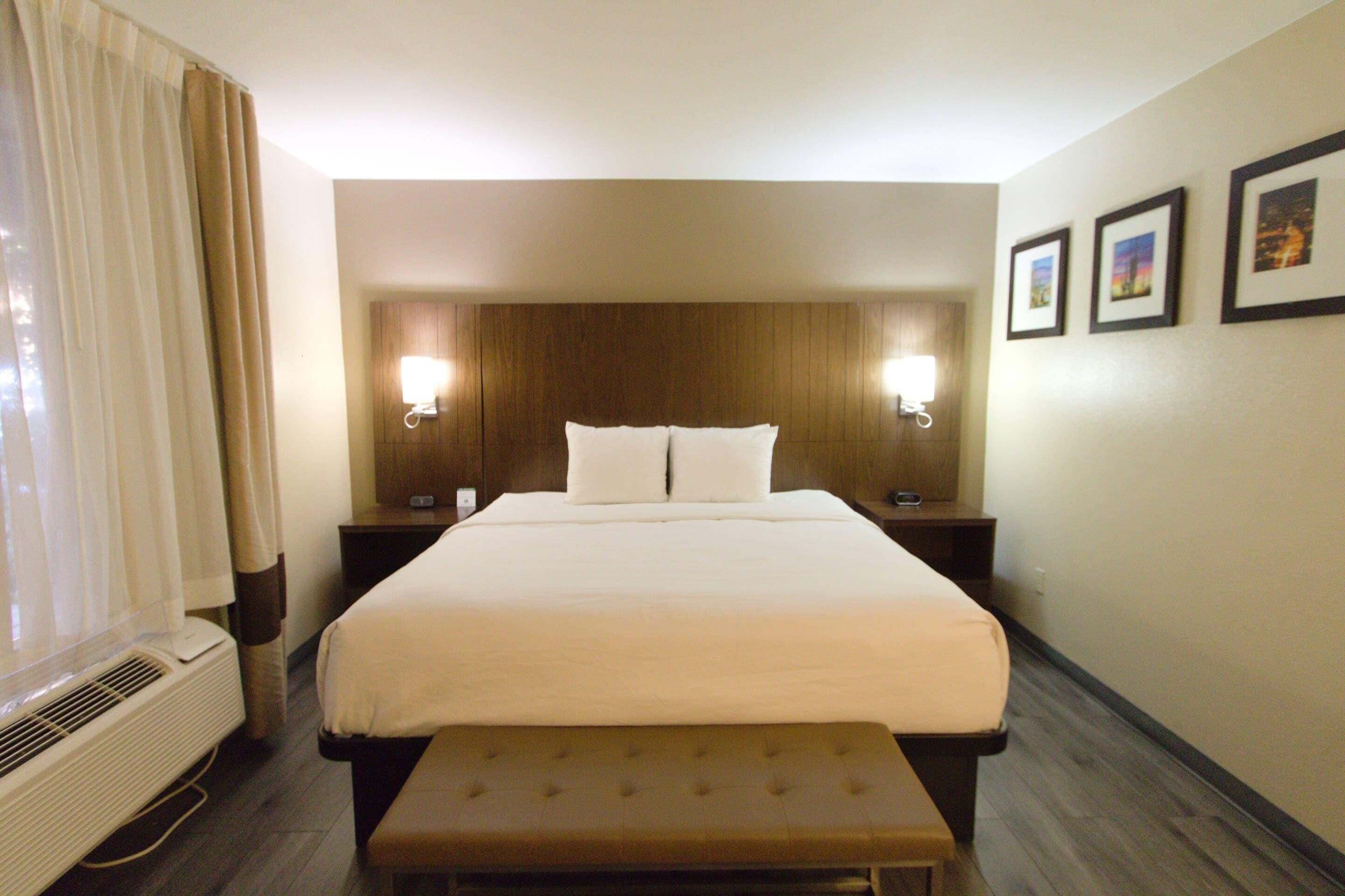 A hotel room with a bed, bedside lamps, a bench, a wall-mounted air conditioner, and framed pictures on the wall.