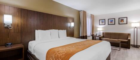 Habitación de hotel con una cama grande, cabecera de madera, mesita de noche con lámpara, sofá y televisor instalado en la pared.