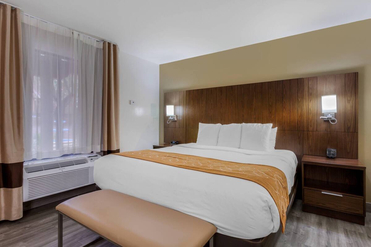 A hotel room with a bed, bedside tables, a bench, a window with curtains, and a wall-mounted air conditioner.