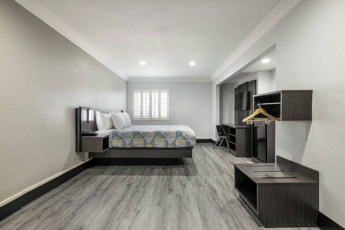 A modern hotel room with a bed, bedside table, wardrobe, and a desk area.
