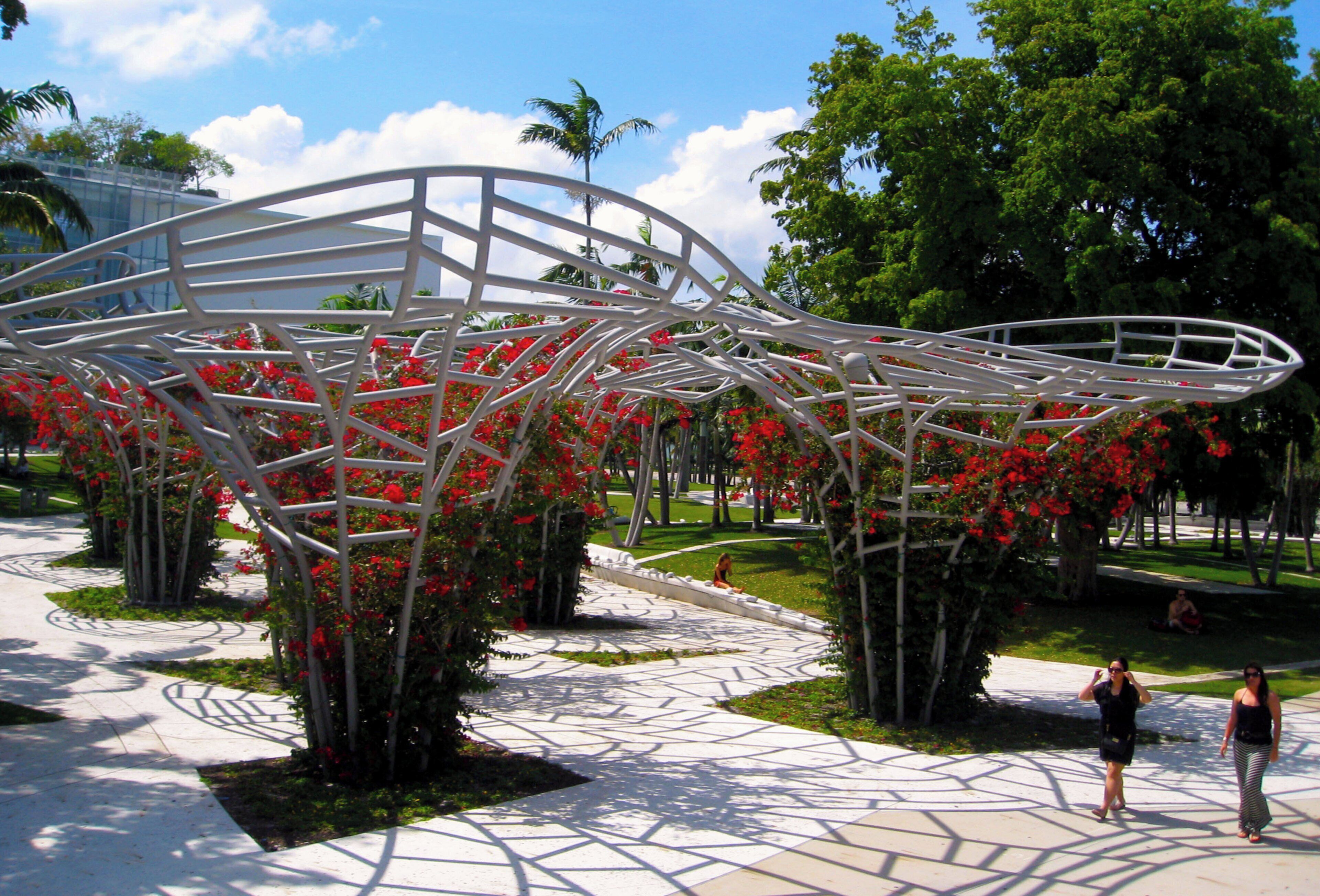 A modern outdoor structure with geometric metal framework, surrounded by red flowers and greenery.