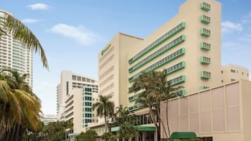 A hotel with a modern design, featuring a prominent green and beige color scheme, a central entrance, and a surrounding palm tree-lined street.