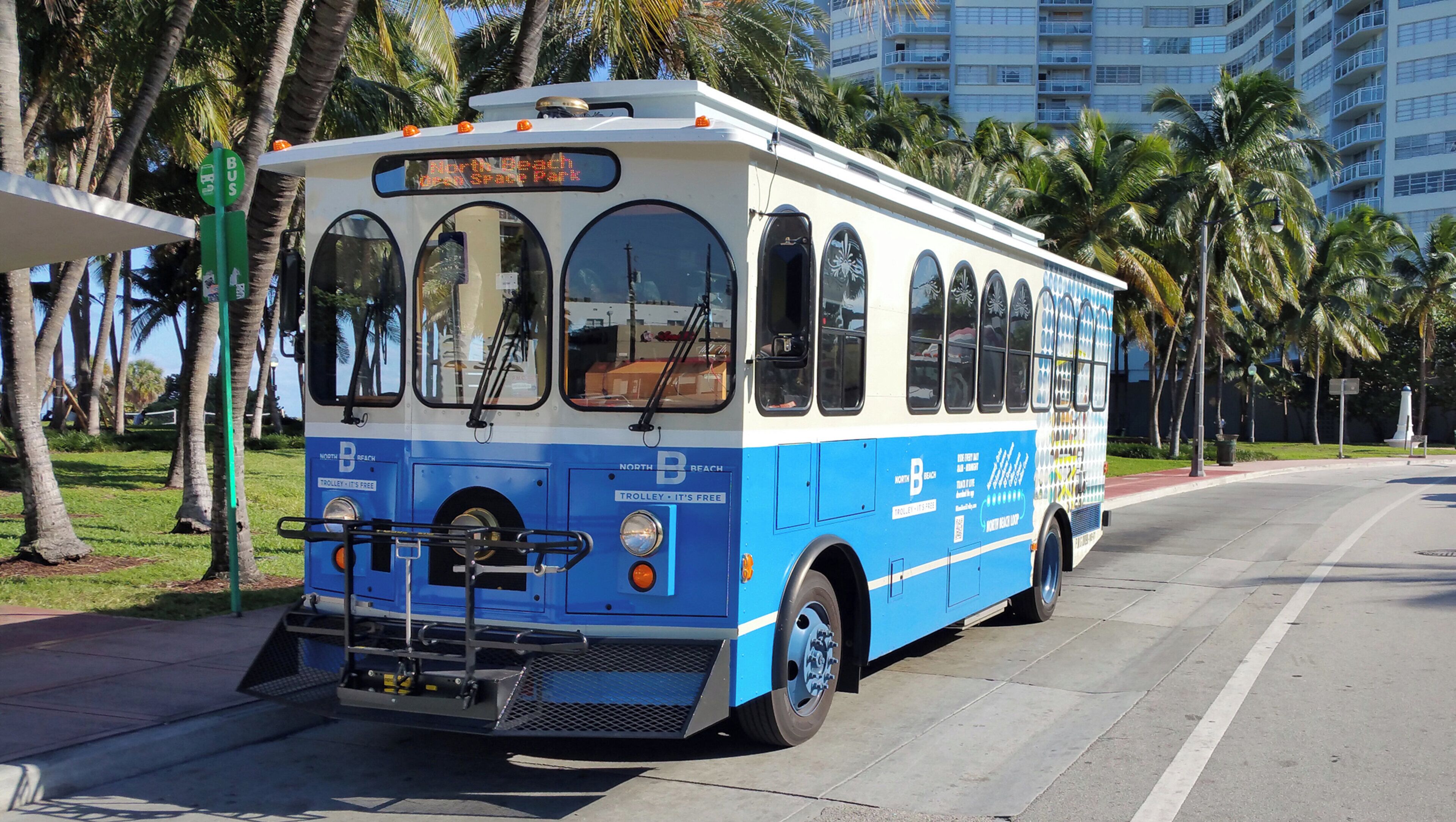 Ein blau-weißer Bus mit der Aufschrift 'North Beach' und 'Trolley – It's Free', geparkt in einer Straße mit Palmen und einem Gebäude im Hintergrund.