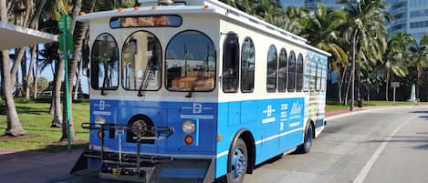Un autobús azul y blanco con el texto 'North Beach' y 'Trolley - It's Free', estacionado en una calle con palmeras y un edificio al fondo.