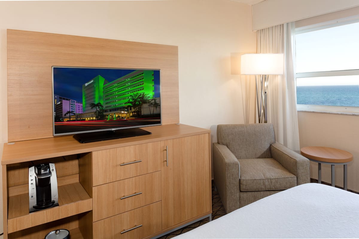 A hotel room with a flat-screen TV mounted on a wooden cabinet, a grey armchair, a small round table, and a bed with white bedding.