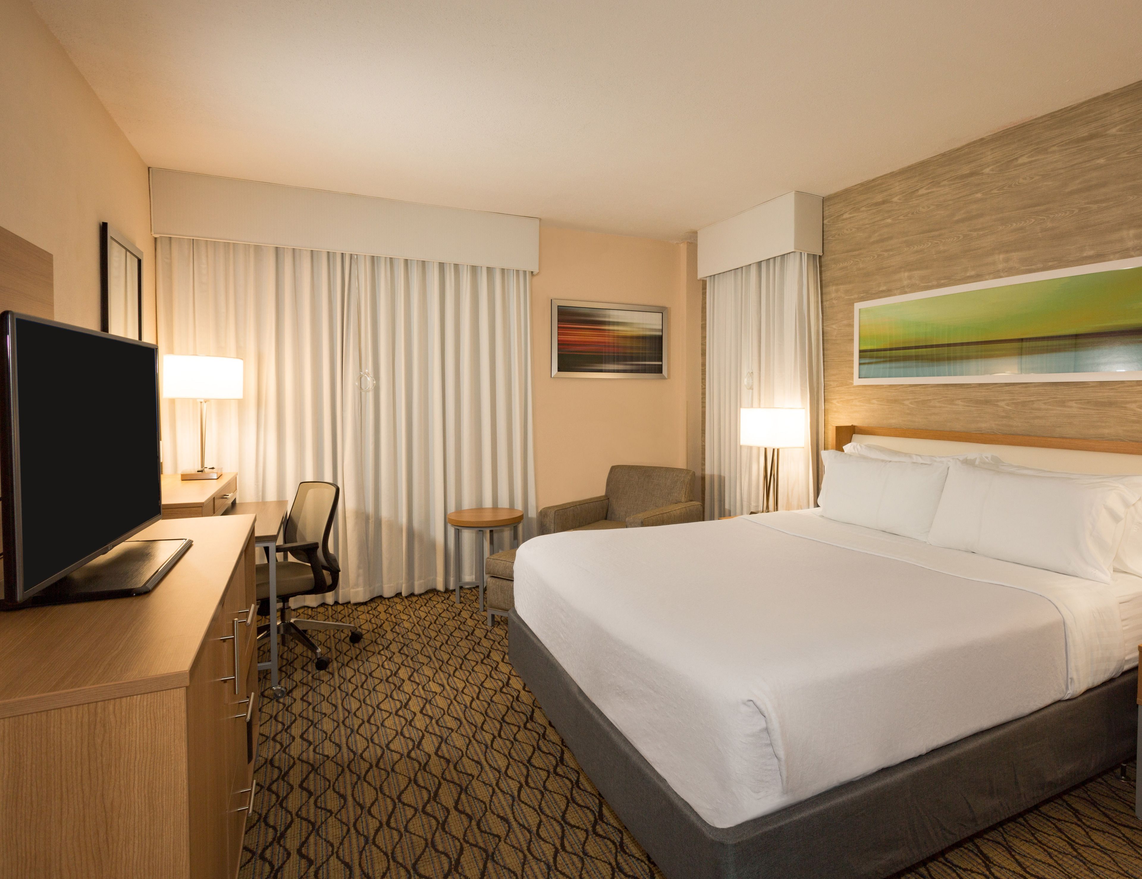 Una habitación de hotel con cama, escritorio con televisor, silla, mesita y un cuadro en la pared.