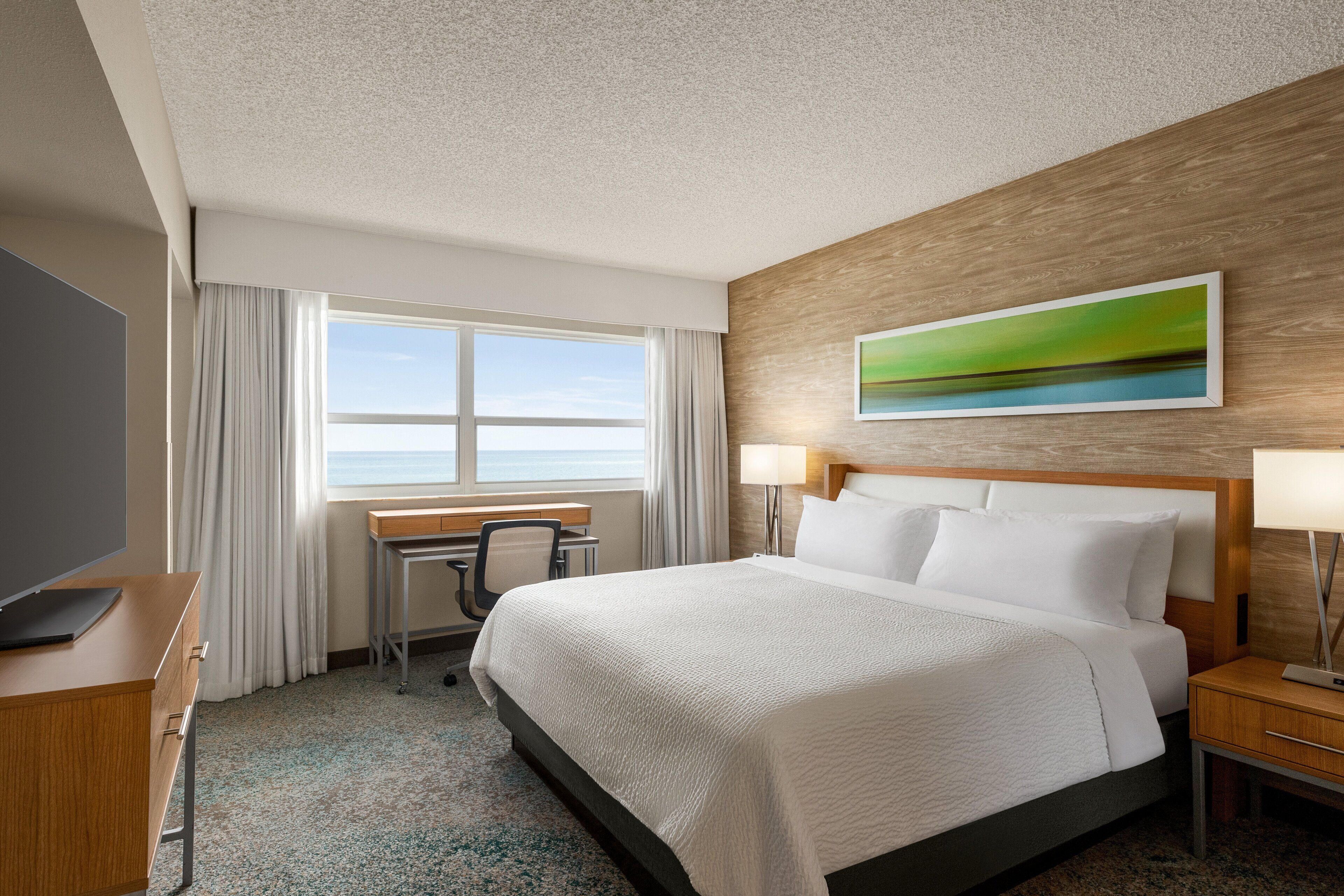 A hotel room with a bed, desk, chair, and a painting on the wall.