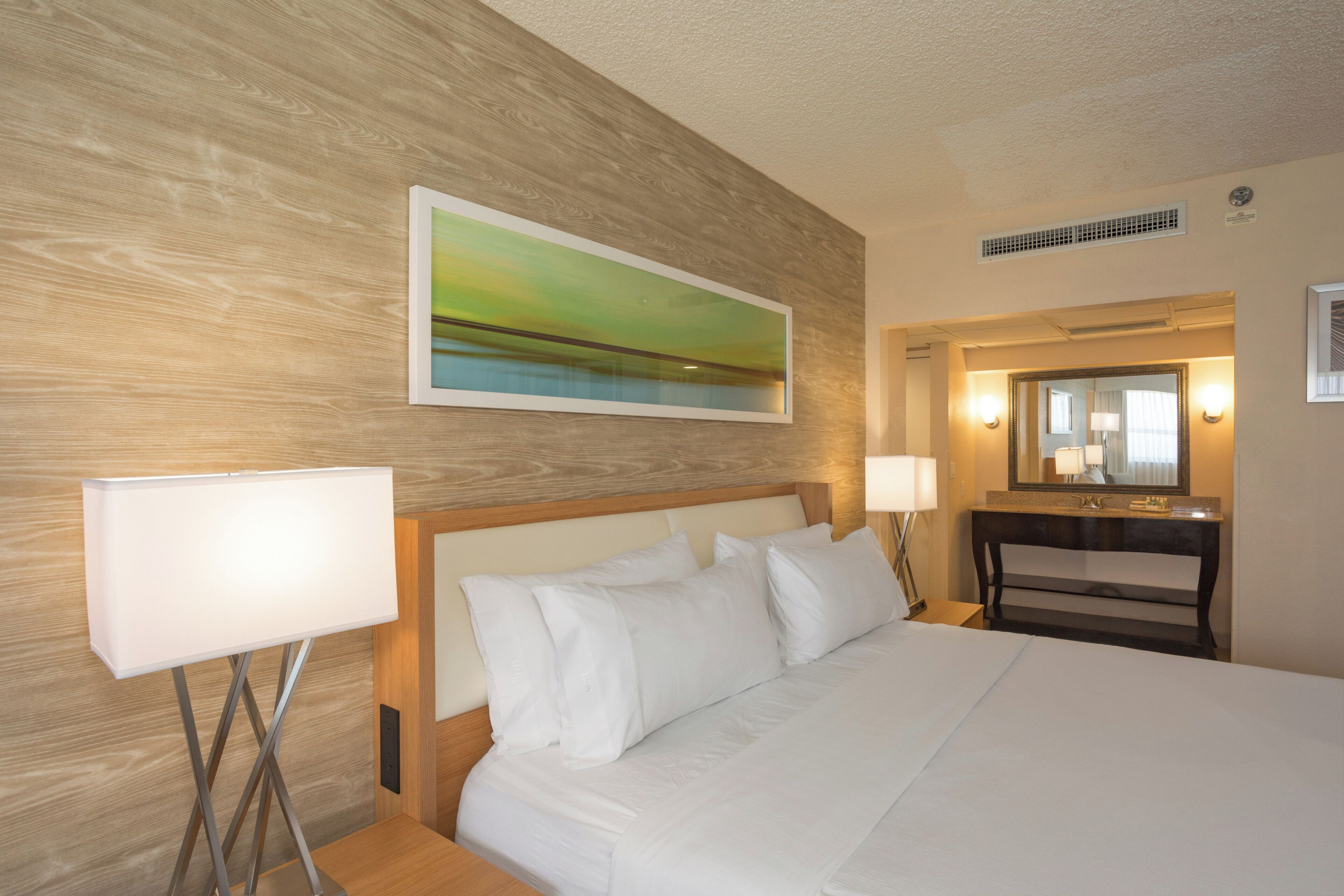 A hotel room with a bed, a nightstand, a lamp, a painting, a bathroom with a mirror, and a wall-mounted vent.