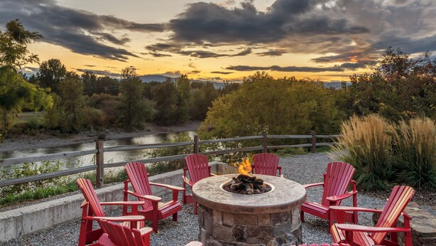 A circular stone fire pit with red Adirondack chairs around it, set on a gravel area with a river and forest in the background.