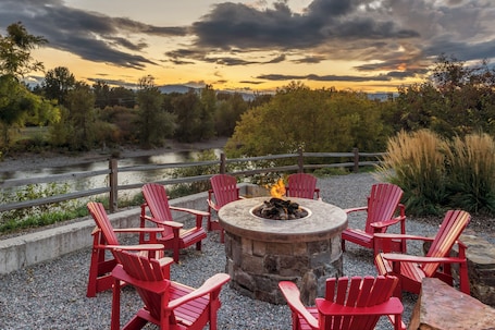 A circular stone fire pit with red Adirondack chairs around it, set on a gravel area with a river and forest in the background.