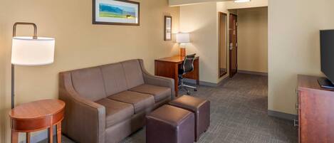 A hotel room with a sofa, two ottomans, a desk with a chair, a lamp, and a television.