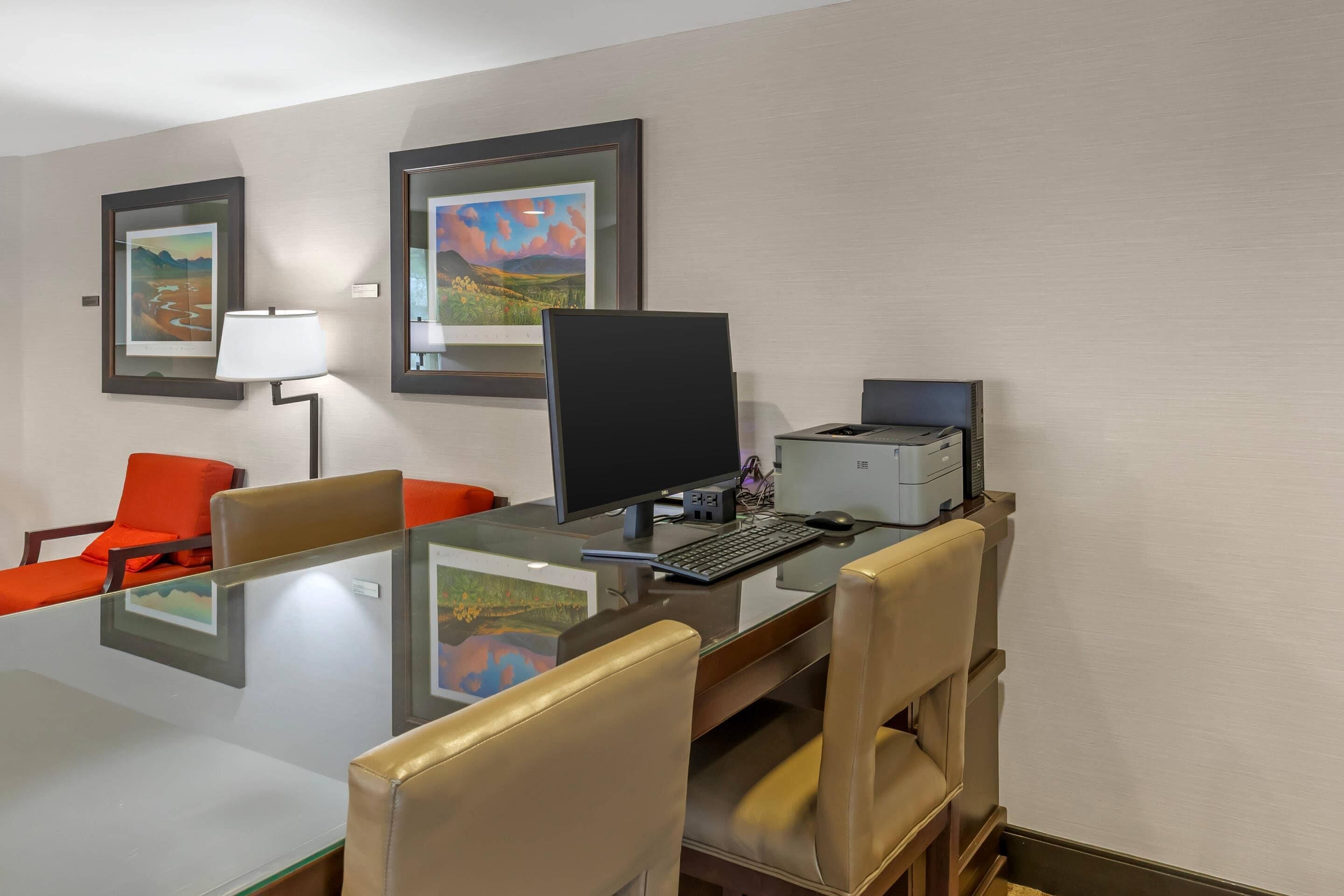 Habitación de hotel con una mesa de conferencias de vidrio, sillas de cuero, una computadora de escritorio, una impresora y obras de arte enmarcadas en las paredes.
