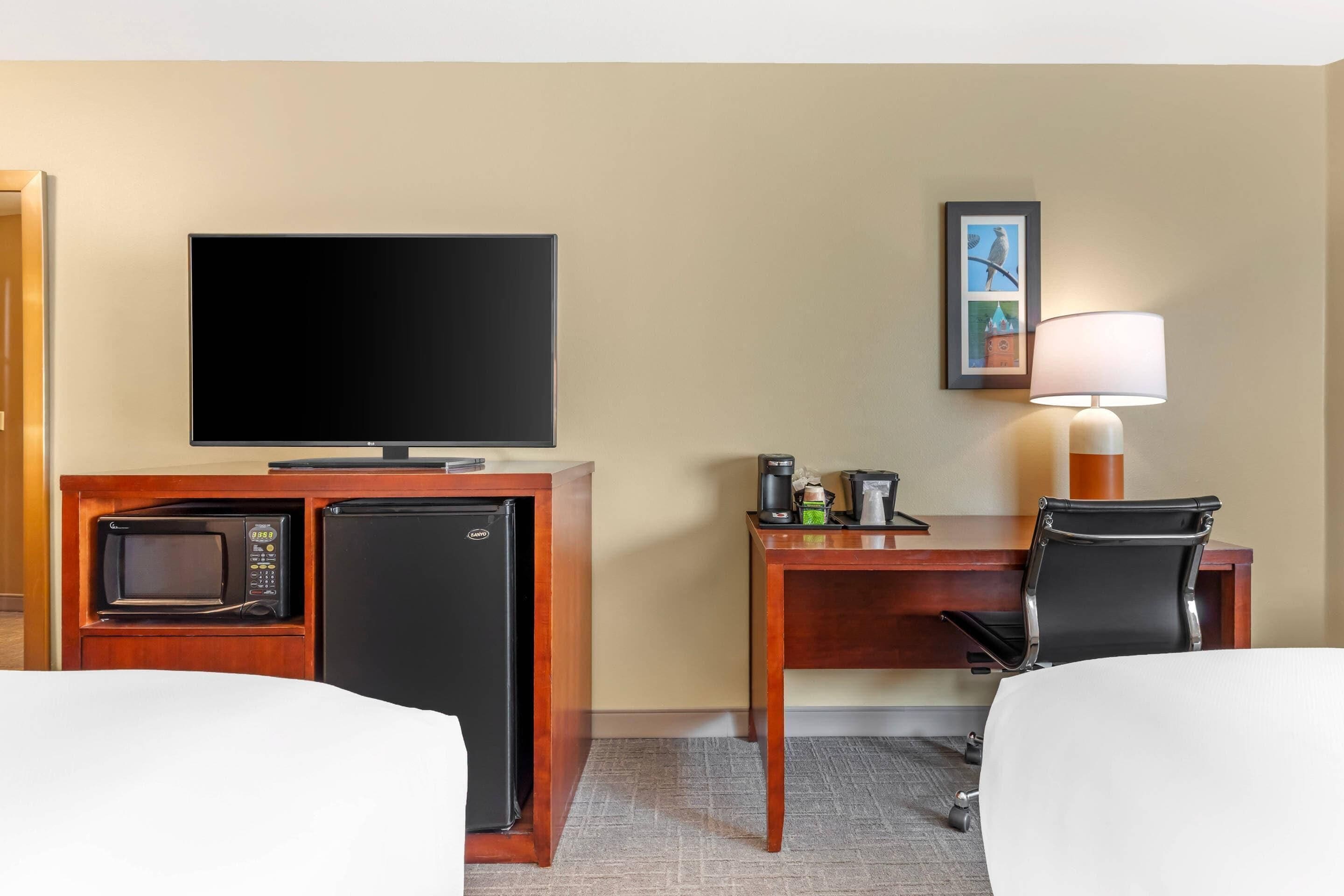 Ein Hotelzimmer mit Fernseher, Schreibtisch mit Lampe, Mikrowelle, Kühlschrank und einem Sessel.