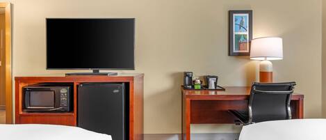 A hotel room with a television, a desk with a lamp, a microwave, a refrigerator, and a chair.