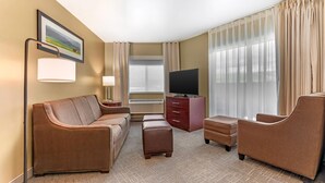 A hotel room with a sofa, ottoman, armchair, TV, and a large window with curtains.