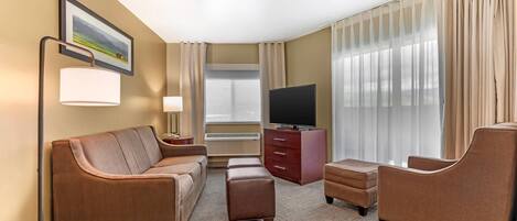 Ein Hotelzimmer mit Sofa, Hocker, Ohrensessel, Fernseher und einem großen Fenster mit Vorhängen.