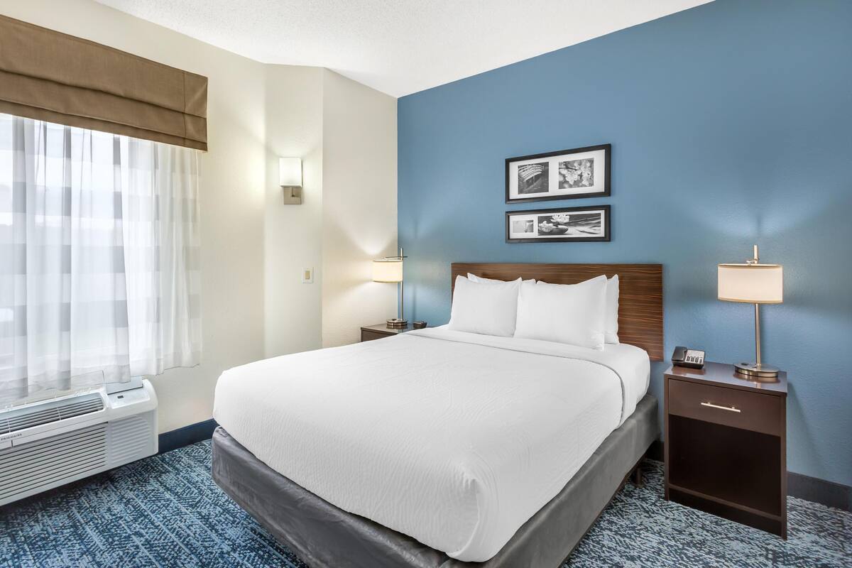 A hotel room with a large bed, two bedside tables with lamps, a wall-mounted air conditioner, and a window with sheer curtains.