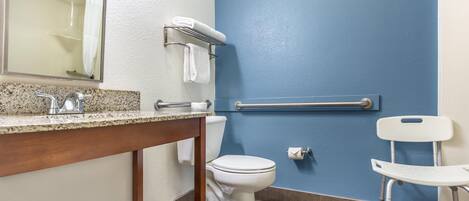 A bathroom with a white toilet, a sink with a granite countertop, a mirror, a towel rack, a handrail, and a white chair.